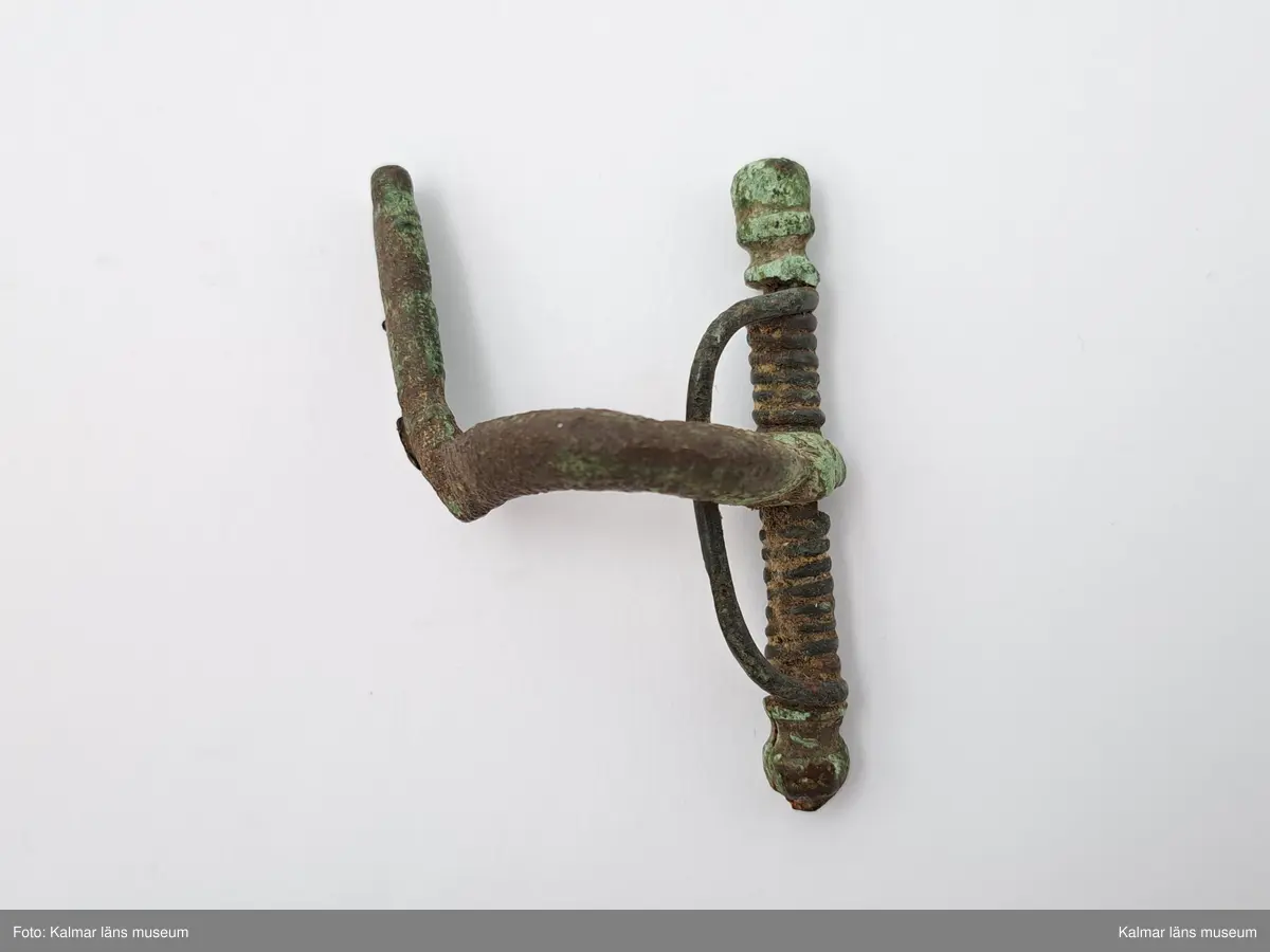 KLM 1248. Fibula, armborstfibula. Fotstycket slutar tvärt nedtill. Nålhållaren är böjd vinkelrätt åt sidan. Tvärstång med olikformade knoppar.