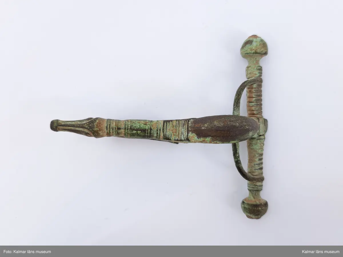 KLM 1458. Fibula, armborstfibula. Gjuten av brons. Fotstycket avslutas med ett djurhuvud och nålhållaren undertill är helt öppen. Själva nålen saknas. Tvärstång med ändknoppar.
