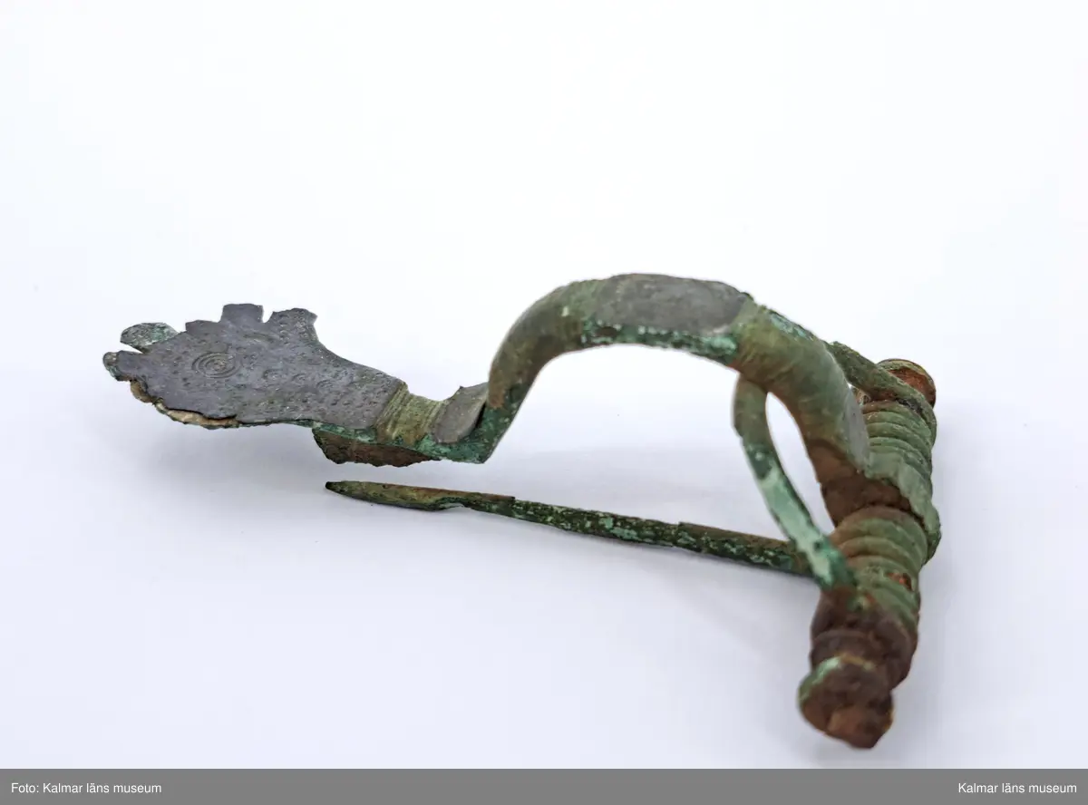 KLM 1979. Fibula, armborstfibula. Gjuten av brons. Det breda fotstycket och den plana bågens rygg är silverbelagd. Fotstycke och båge ornerad med åsar, punkter och cirklar. Tvärstång med ändknoppar, den ena skadad.