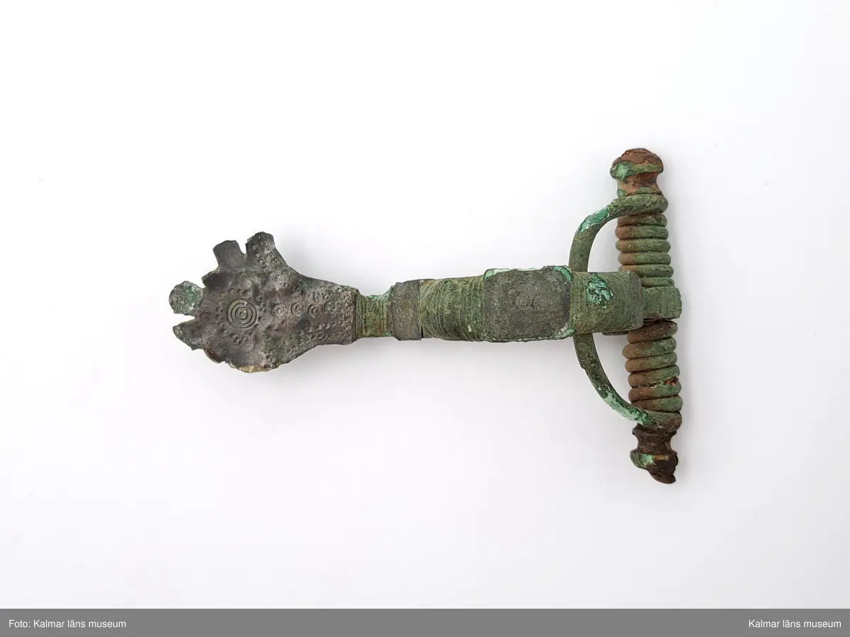 KLM 1979. Fibula, armborstfibula. Gjuten av brons. Det breda fotstycket och den plana bågens rygg är silverbelagd. Fotstycke och båge ornerad med åsar, punkter och cirklar. Tvärstång med ändknoppar, den ena skadad.