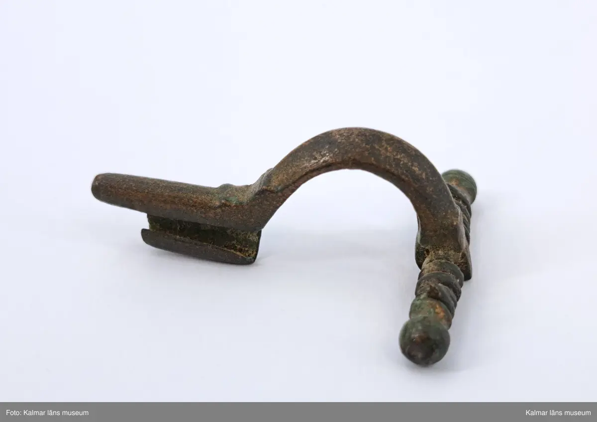 KLM 2938. Fibula, armborstfibula. Gjuten av brons. Tvärstången slutar i runda knoppar, spiralens sträng saknas. Bågen svagt ryggad och nålhållaren är något kortare än fotstycket.