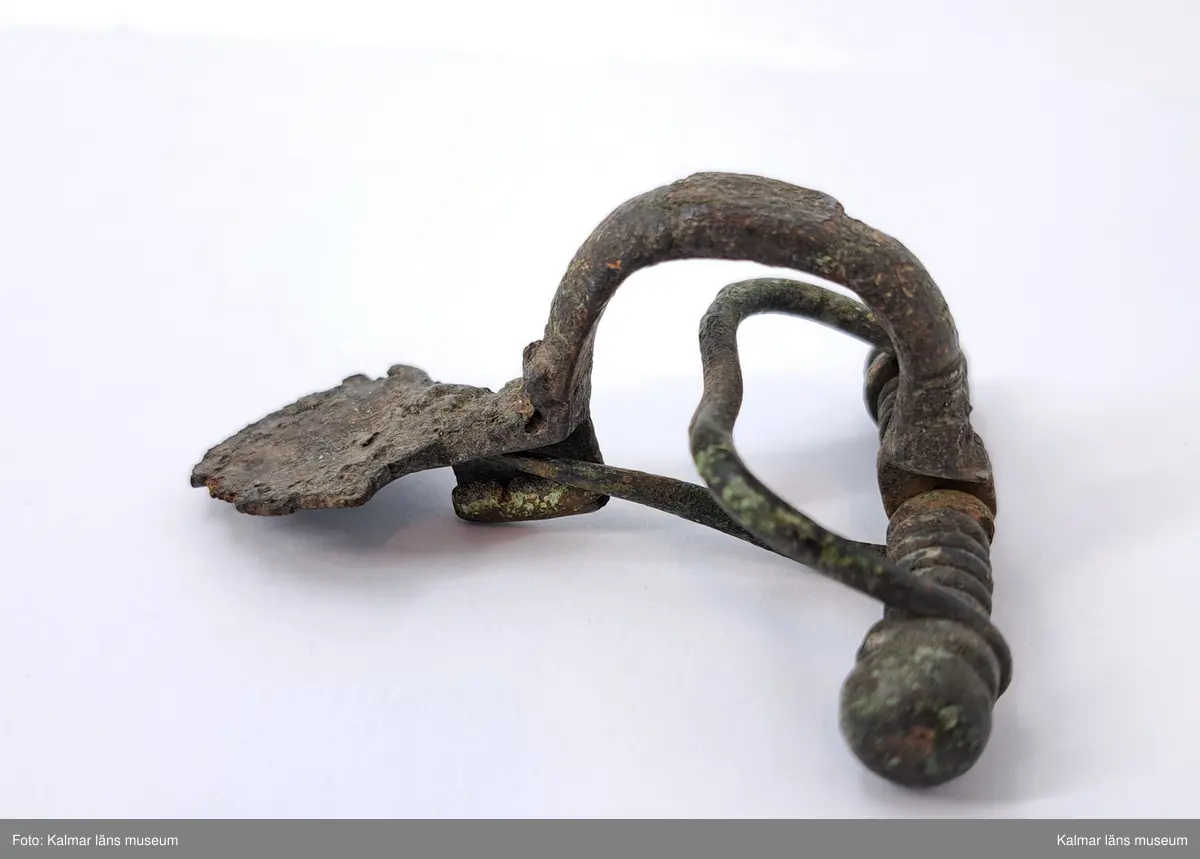 KLM 2165. Fibula, armborstfibula. Gjuten av brons med försilvring. Fotstycket är brett och belagt med silver. Bågens rygg plan och silverbelagd. Tvärstång med ändknoppar.