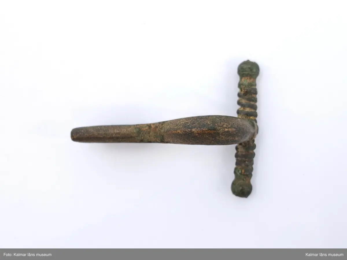 KLM 2938. Fibula, armborstfibula. Gjuten av brons. Tvärstången slutar i runda knoppar, spiralens sträng saknas. Bågen svagt ryggad och nålhållaren är något kortare än fotstycket.