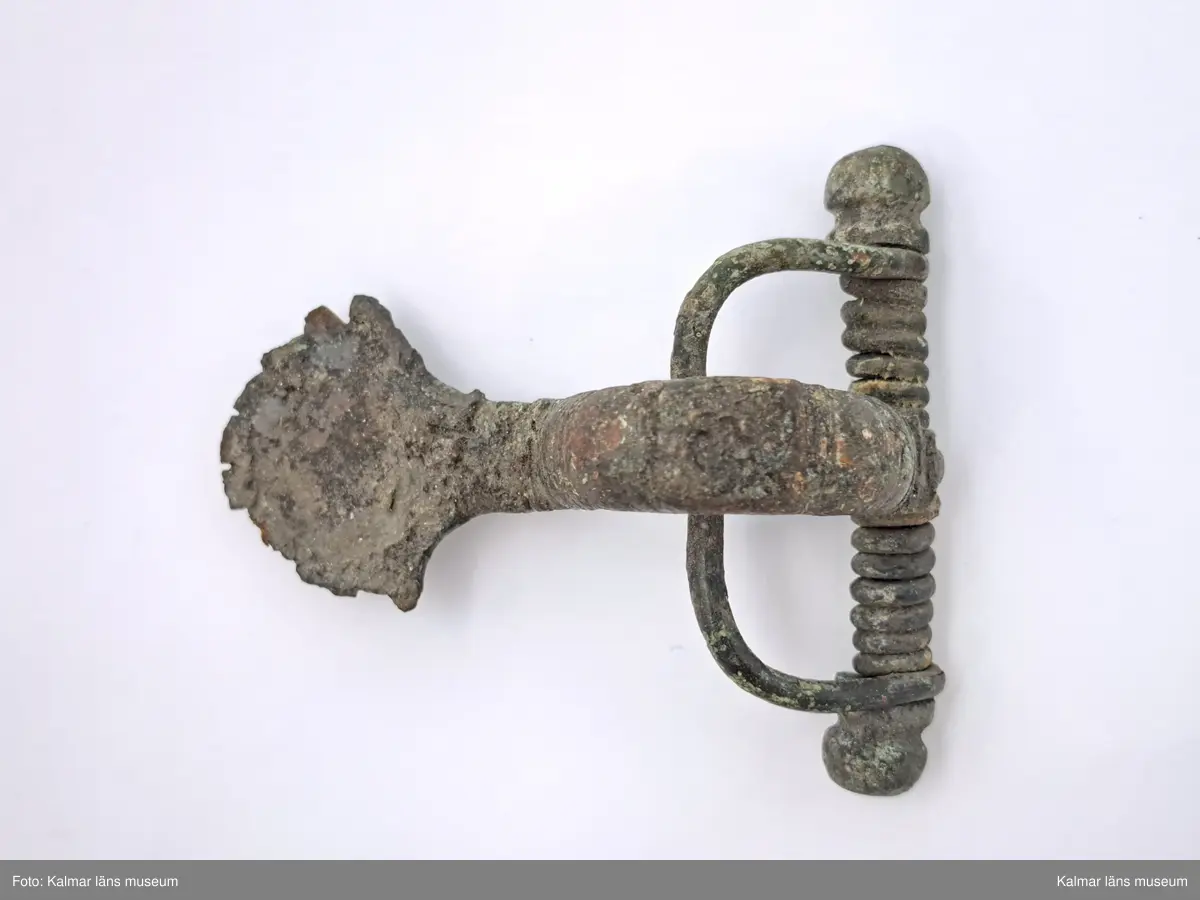 KLM 2165. Fibula, armborstfibula. Gjuten av brons med försilvring. Fotstycket är brett och belagt med silver. Bågens rygg plan och silverbelagd. Tvärstång med ändknoppar.
