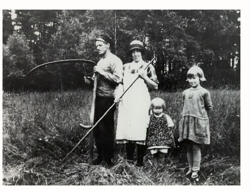 "Skördearbete i Kråkarp omkring 1940." Bilden föreställer förmodligen man och hustru samt två döttrar. Mannen bryner lien och kvinnan räfsar.