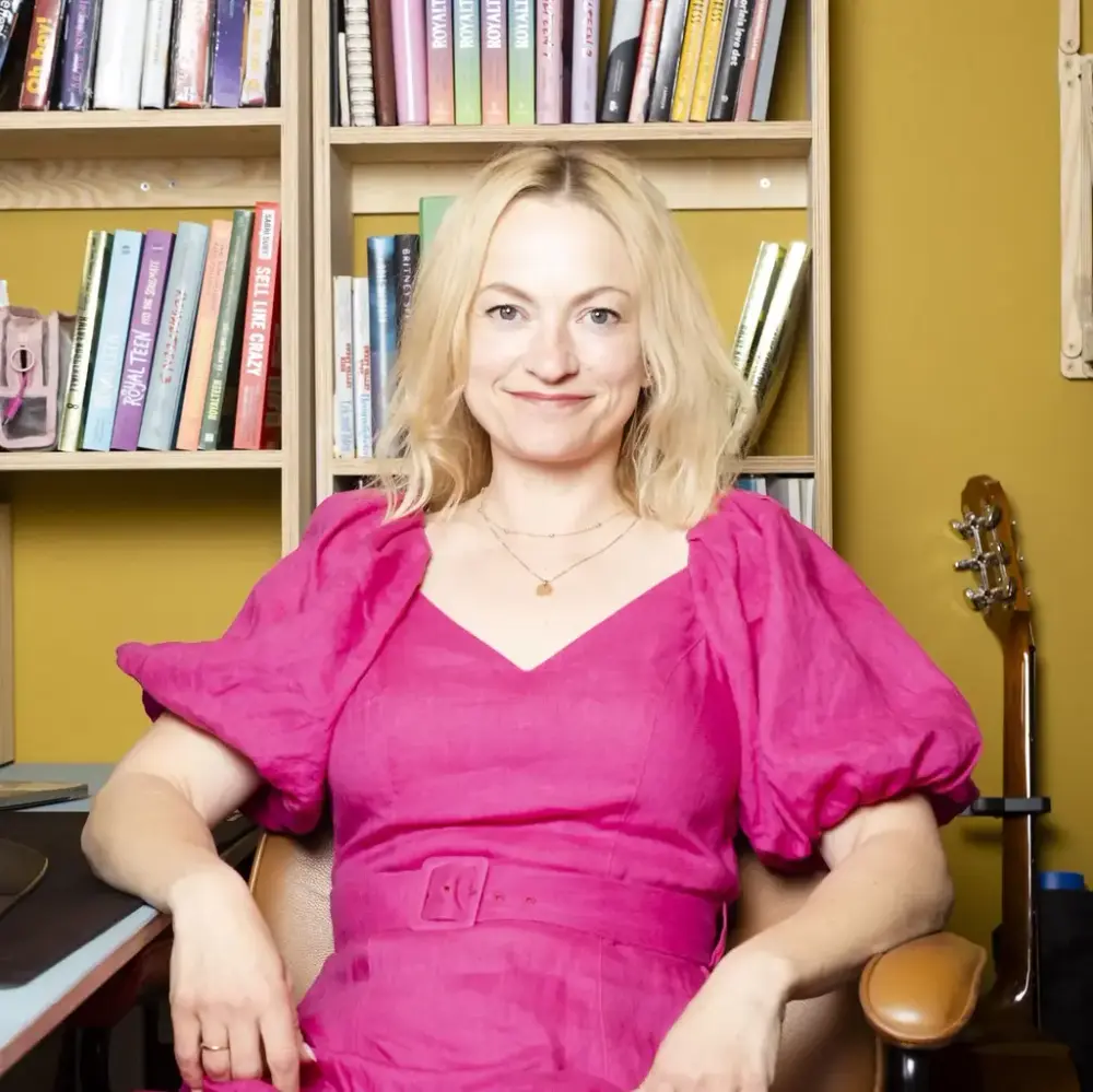 Anne Gunn Halvorsen i rosa kjole sitjande framom ei bokhylle