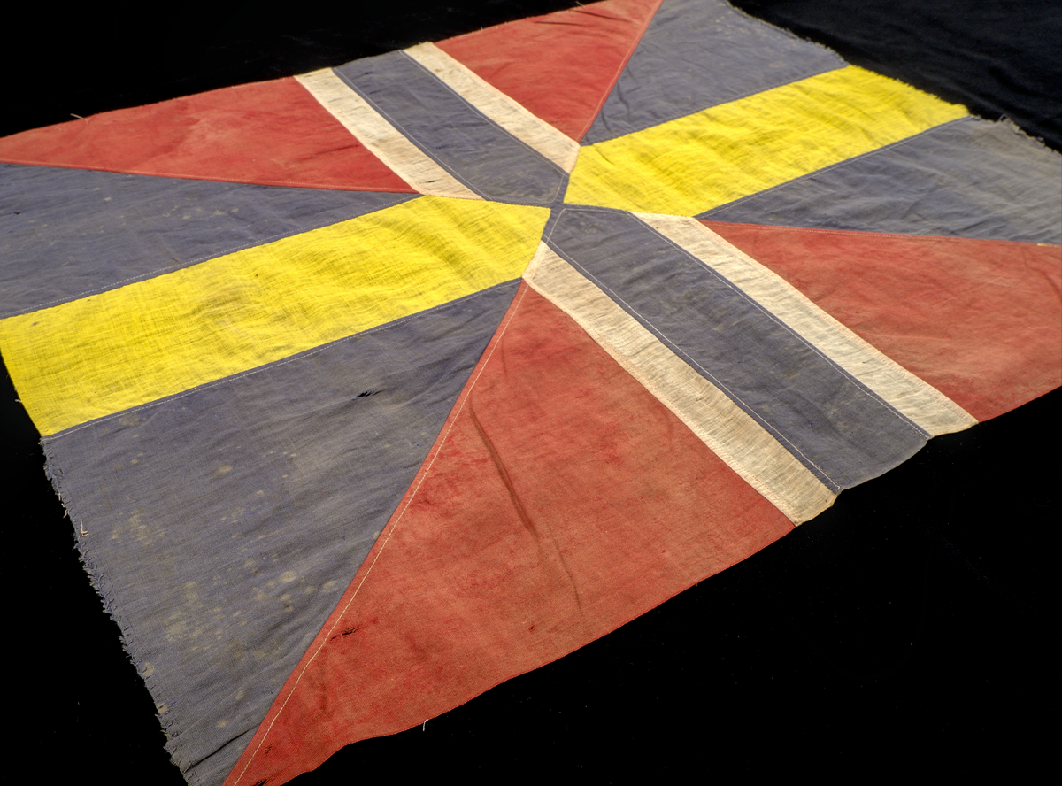 Unionsmärke från svensk-norsk flagga (1844–1905)

Rektangulärt tygmärke, sammansatt av flera tygstycken i tuskaftad bomull, som tillsammans bildar ett korslagt mönster av färgfält i rött, blått, vitt och gult. Styckena är sammanfogade med maskinsydda sömmar i vit bomullstråd.

Föremålet är skört, blekt och delvis nedfläckat med färg. Kaststygnen längs kanterna tillkom i samband med konservering år 2025.