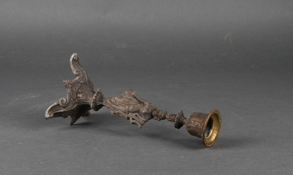 Lysestake av metall. Den har skaft formet som en kvinne som holder en lyre i venstre hånd. Hun står på et trefotet fotstykke. Øverst med pipe formet som en urne med perlebord langs kanten. Hele overflaten på lysestaken har støpt dekor.