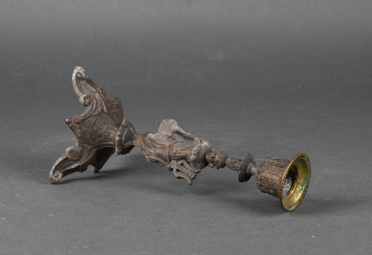 Lysestake av metall. Den har skaft formet som en kvinne som holder en lyre i venstre hånd. Hun står på et trefotet fotstykke. Øverst med pipe formet som en urne med perlebord langs kanten. Hele overflaten på lysestaken har støpt dekor.