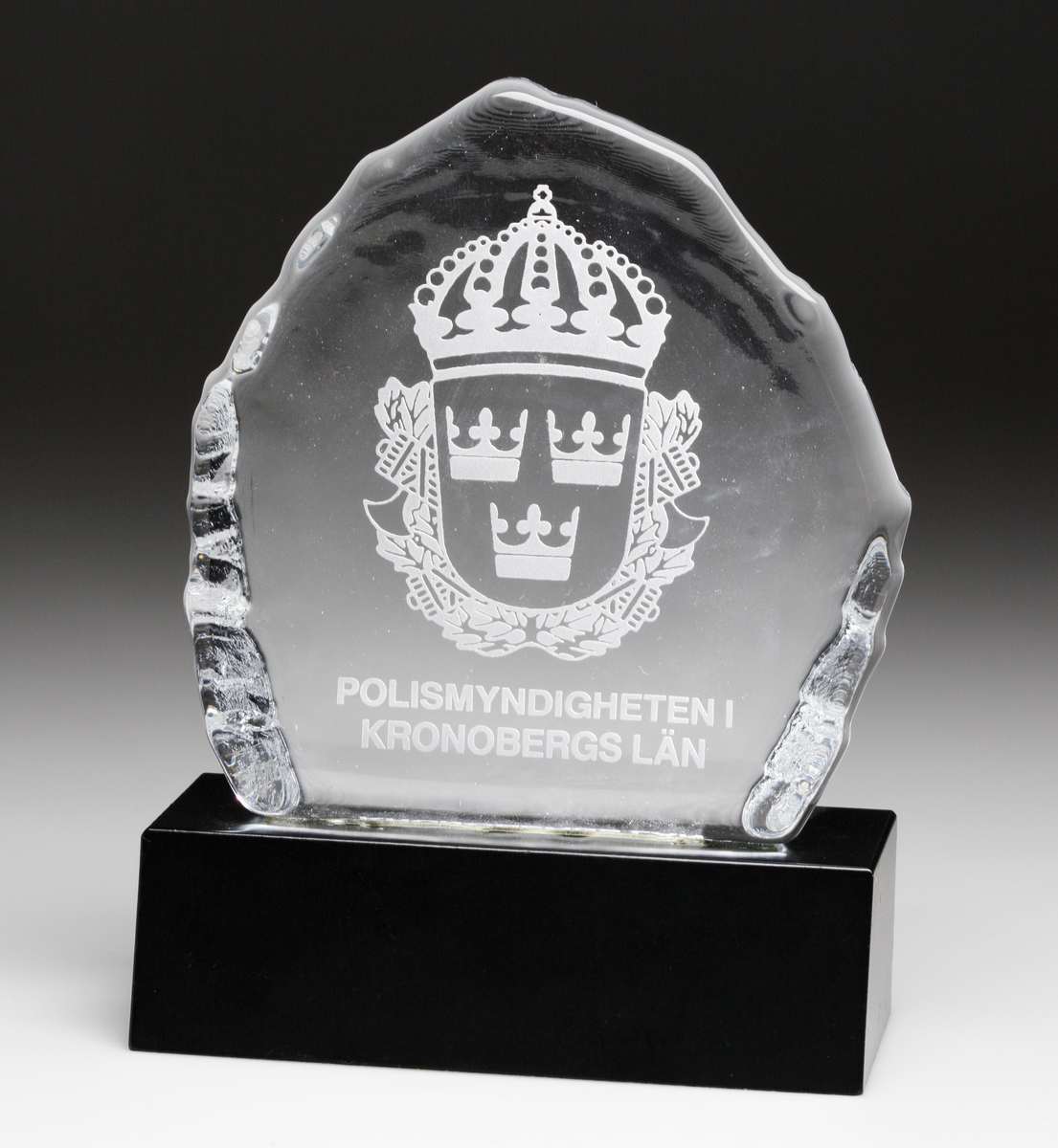 Glasplakett på svart träsockel. Etsat motiv bestående av svenska polisens vapenskjöld, samt texten "POLISMYNDIGHETEN I KRONOBERGS LÄN".