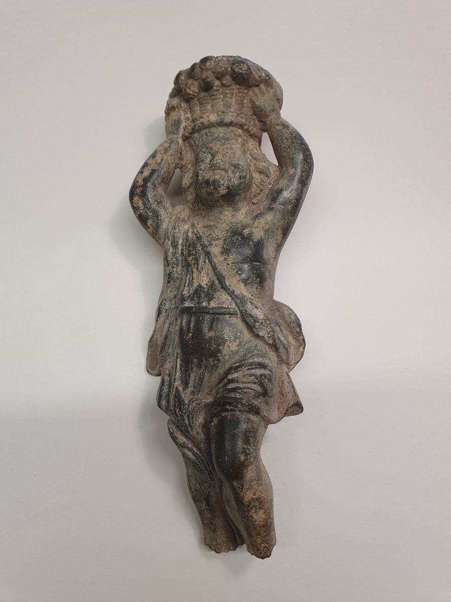 Föremål av brons. Ljushållare i form av putti. Sannolikt del av ljusstake, kandelaber eller liknande. Figuren har armarna uppåtsträckta över huvudet och håller i en korg med frukter. Klädedräkten påminner om en toga med mantel och livrem. Alternativt hänger en väska eller påse över axeln. Mellan benen slingrar en liljeväxt. Figuren är avbruten vid fötterna. Sannolik datering 1600-1700-tal.