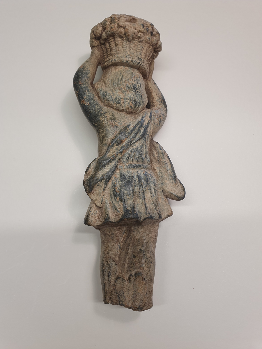 Föremål av brons. Ljushållare i form av putti. Sannolikt del av ljusstake, kandelaber eller liknande. Figuren har armarna uppåtsträckta över huvudet och håller i en korg med frukter. Klädedräkten påminner om en toga med mantel och livrem. Alternativt hänger en väska eller påse över axeln. Mellan benen slingrar en liljeväxt. Figuren är avbruten vid fötterna. Sannolik datering 1600-1700-tal.