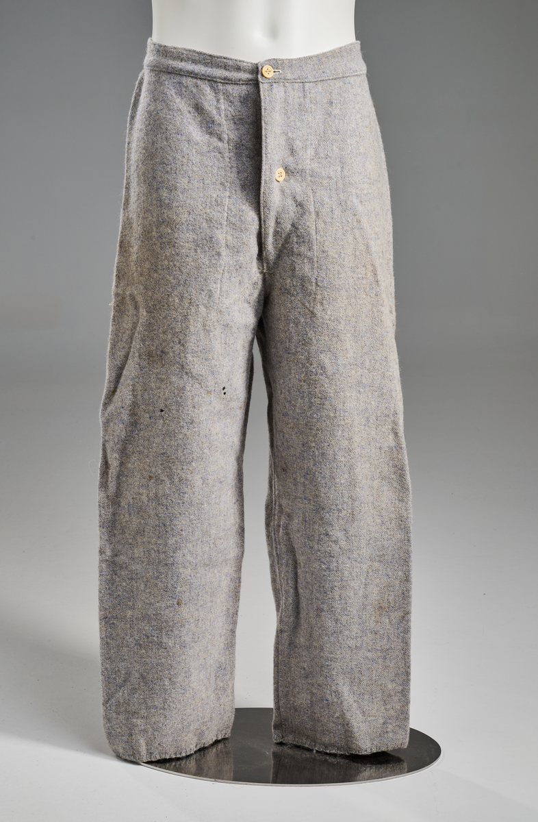 Lyseblå/grå vadmelsbukse, kypertvevd. Gylf med dobbelt stoff og to hornknapper, hvite, to knapphull. Bred linning med påsydd lyseblått bomullsstoff på innsiden. Beina smalner skrått, kantet med tungesting nederst. Tre legg på hver side av midtsømmen bak.