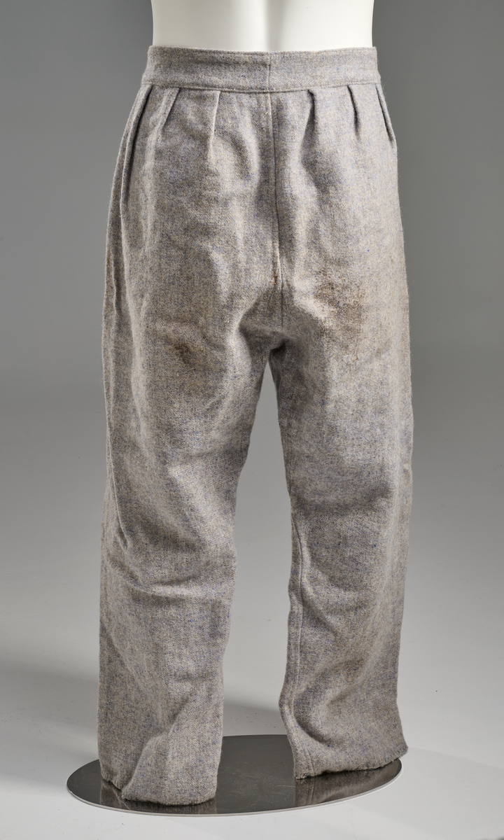 Lyseblå/grå vadmelsbukse, kypertvevd. Gylf med dobbelt stoff og to hornknapper, hvite, to knapphull. Bred linning med påsydd lyseblått bomullsstoff på innsiden. Beina smalner skrått, kantet med tungesting nederst. Tre legg på hver side av midtsømmen bak.