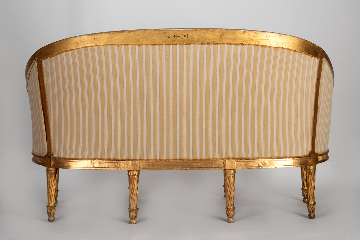 Sofa med langstrakt, oval sargramme med forsenket, riflet midtparti og i fronten et symmetrisk skåret grenornament. Ved festet for hvert av de fire rette frambeina er det en rektangulær plate med rosett. Beina har skråriflet midtparti, øverst krans med "dråper" og nederst skåret løv avsluttet med liten kule. Beina på hver side, fortsetter opp i de runde, kannelerte armstøttene. Den høye, buede ryggrammen har samme dekorative utforming som sargen og hulkil langs oversiden. Den avsluttes i korte, svungne armlener med opprulling på endene og skåret løv på oversiden. Stoppet sete og rygg, samt løs, stoppet pute. Sofaen er trukket med lyst, beige stoff med gullinnvikning og blomster. Alt treverk er forgylt, også på baksiden.