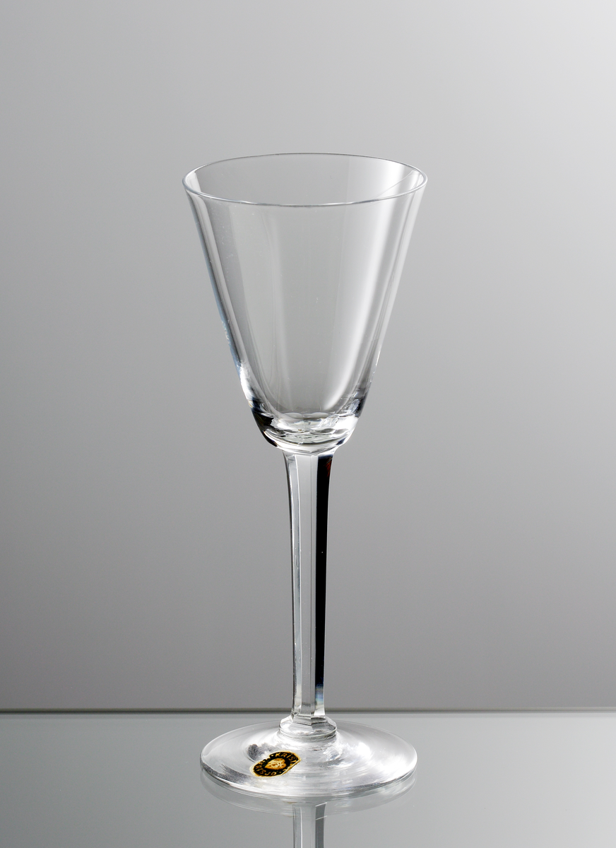 Brännvinsglas med konad kupa och facettslipat högt ben. Rund etikett med blå botten och gul text KRISTALL HIERTSJÖ.