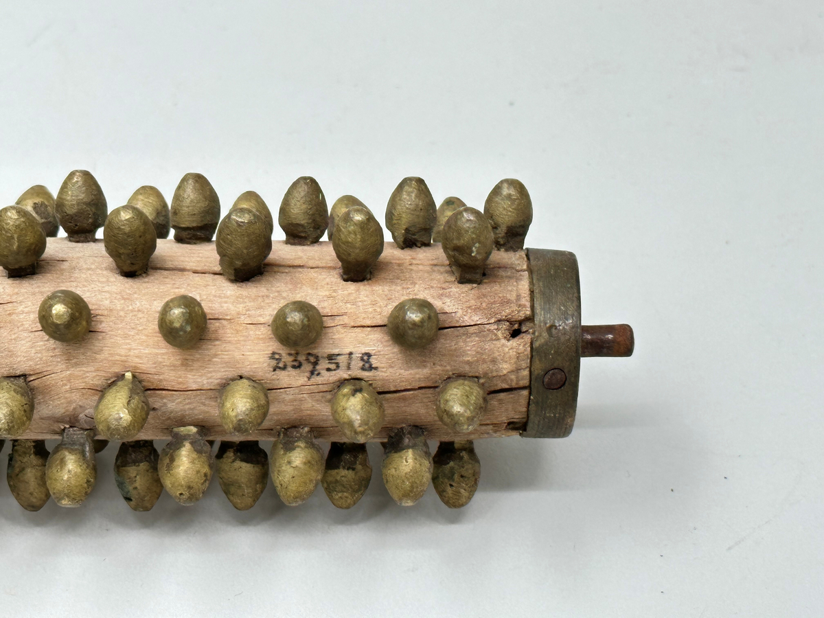 Rullnagg, för spisbröd. Rader av mässingsnaggar, trärulle som sitter i järnfattning, vilket har svarvat träskaft. Trasig, har lossnat från skaftet. Sprucken. 