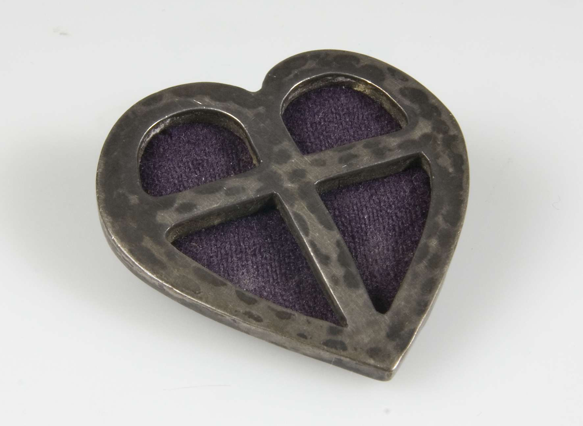 Brosch av hamrat silver i form av ett hjärta med inskuret kors med violett yllefilt bakom.