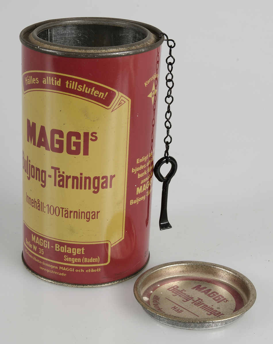 Burk av röd- och gulmålad plåt, med text, bl. a: "MAGGIS  Buljong-Tärningar Innehåll: 100 Tärningar". På burkens överkant hänger en öppnare. Locket kan öppnas och förslutas. Burken är tom.


