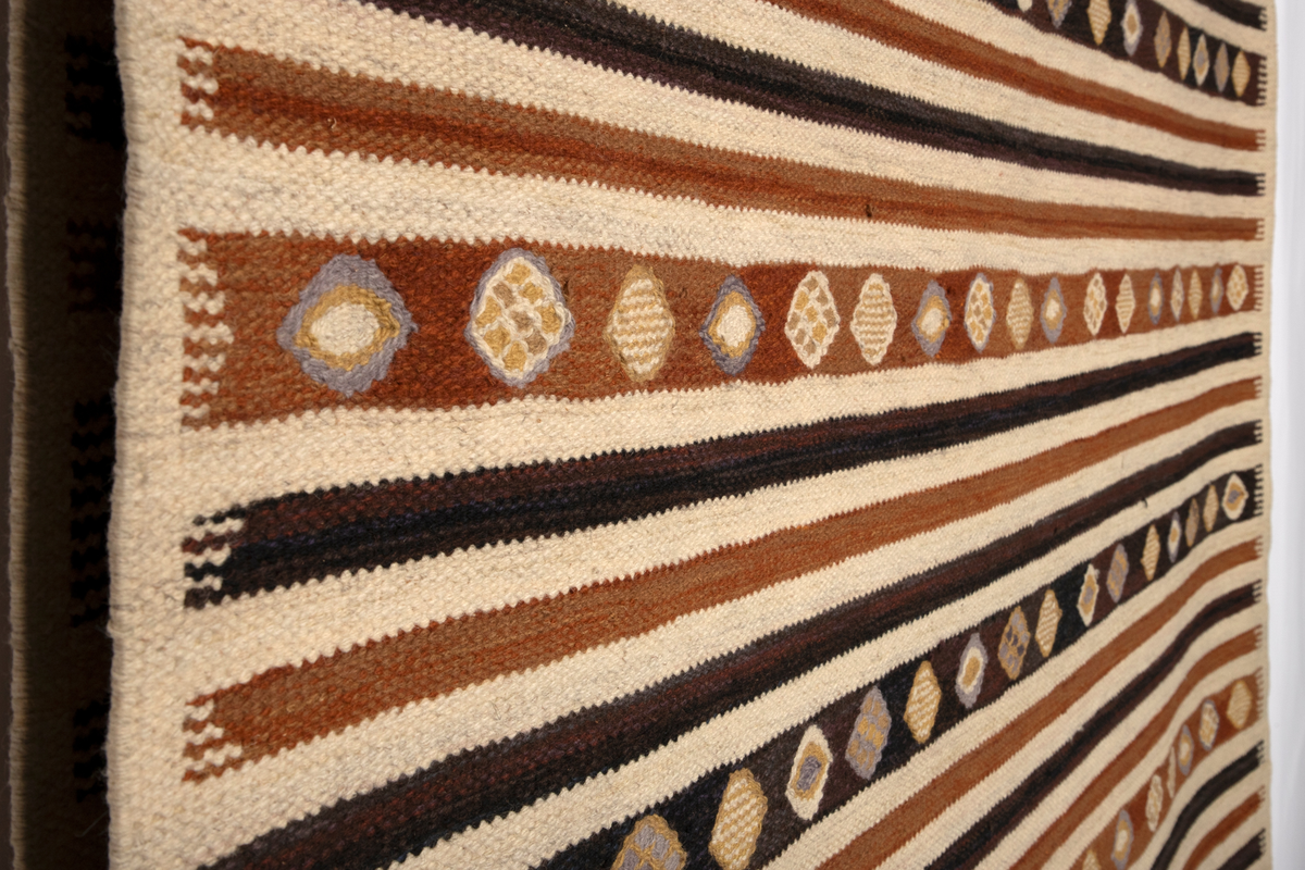 Gulvteppet vevd i röllakanteknikk. Lingarn i renningen og nauthårsgarn i innslaget. Stripet i hvitt, rødbrunt og brunsvart i innlagsretningen, sju smale striper veksler med en bredere stripe hver 25. cm. I de brede stripene er det vevd inn spissovale former med hvitt, lilla og gult fylldekor bestående av avvekslende flekker, striper og spissovaler. På langsidene en 3 cm bred jarekant i hvitt. I teppets kortender er det knyttet frynser av renningen.