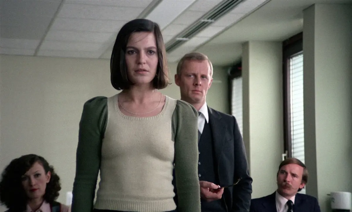 Kvinna som står upp i ett rum med en stående man och två sittande personer i bakgrunden.