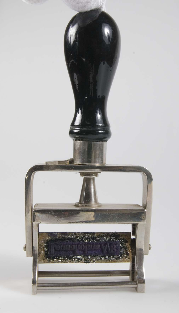 Stämpel av förkromat järn med svart svarvat trähandtag. Stämpel av gummi med texten: "Lennaholm A.B.". Stämpelytan vilar mot en gredelin stämpeldyna och vänds när stämpeln pressas ned. Kan låsas i nedtryckt läge med en spak.
