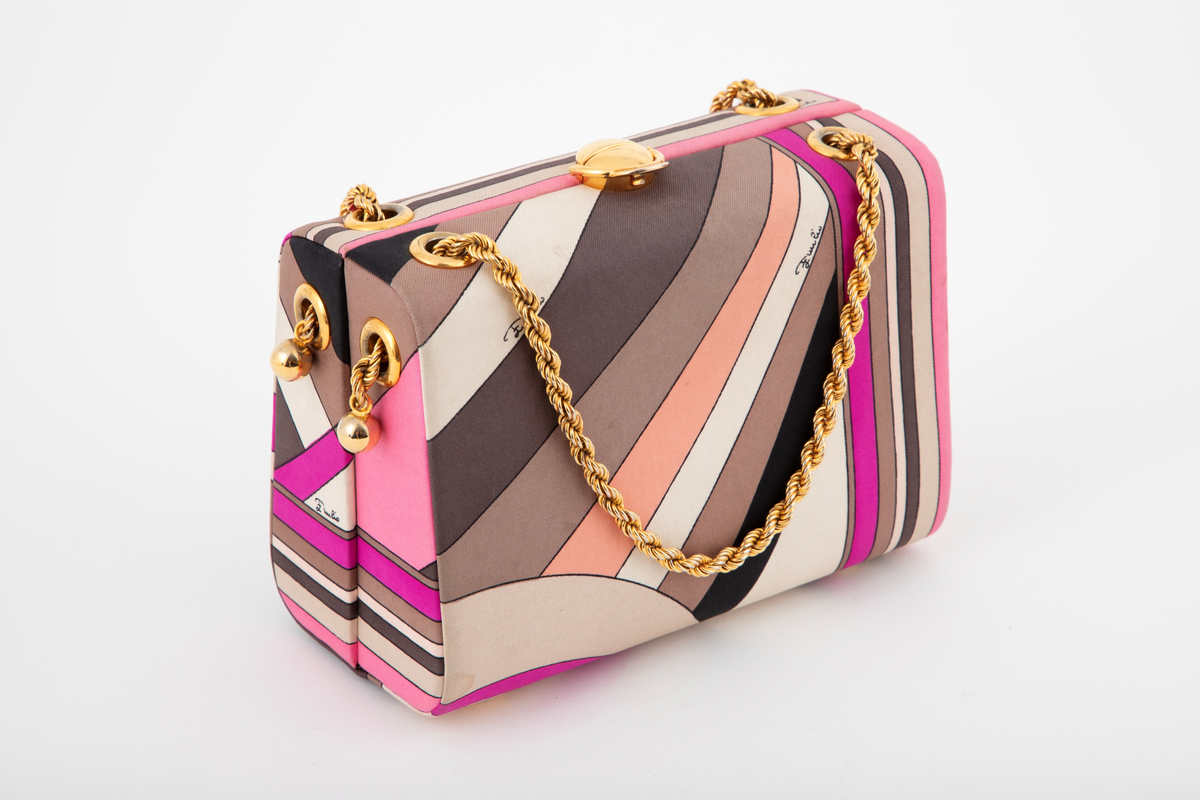 Liten boksveske med ett stort innerrom og et mindre innerrom på bakveggen. Vesken er hard, og må ha en innvendig kjerne av et hardt materiale. Utvendig er den trukket med et silketrykk i sterke rosa nyanser, nonfigurativt, men likevel noe geometrisk av type. Mønstertypen er karakteristisk for designeren Emilio Pucci på sluttet av 1960-tallet og på 1970-tallet. Innvendig kledd med hvitt kidskinn, og stor, båtformet åpning på innerlommen. Innerrommet er fôret med bronsefarget skinn. Knepplås på midten av oversiden og 2 bærelenker, tredd gjennom kraftige maljer på toppen og øverst på kortsidene. De tvunne "gullenkene" holdes på plass av små kuler i endene. På undersiden, 4 små halvkuleformede ben av samme metall.