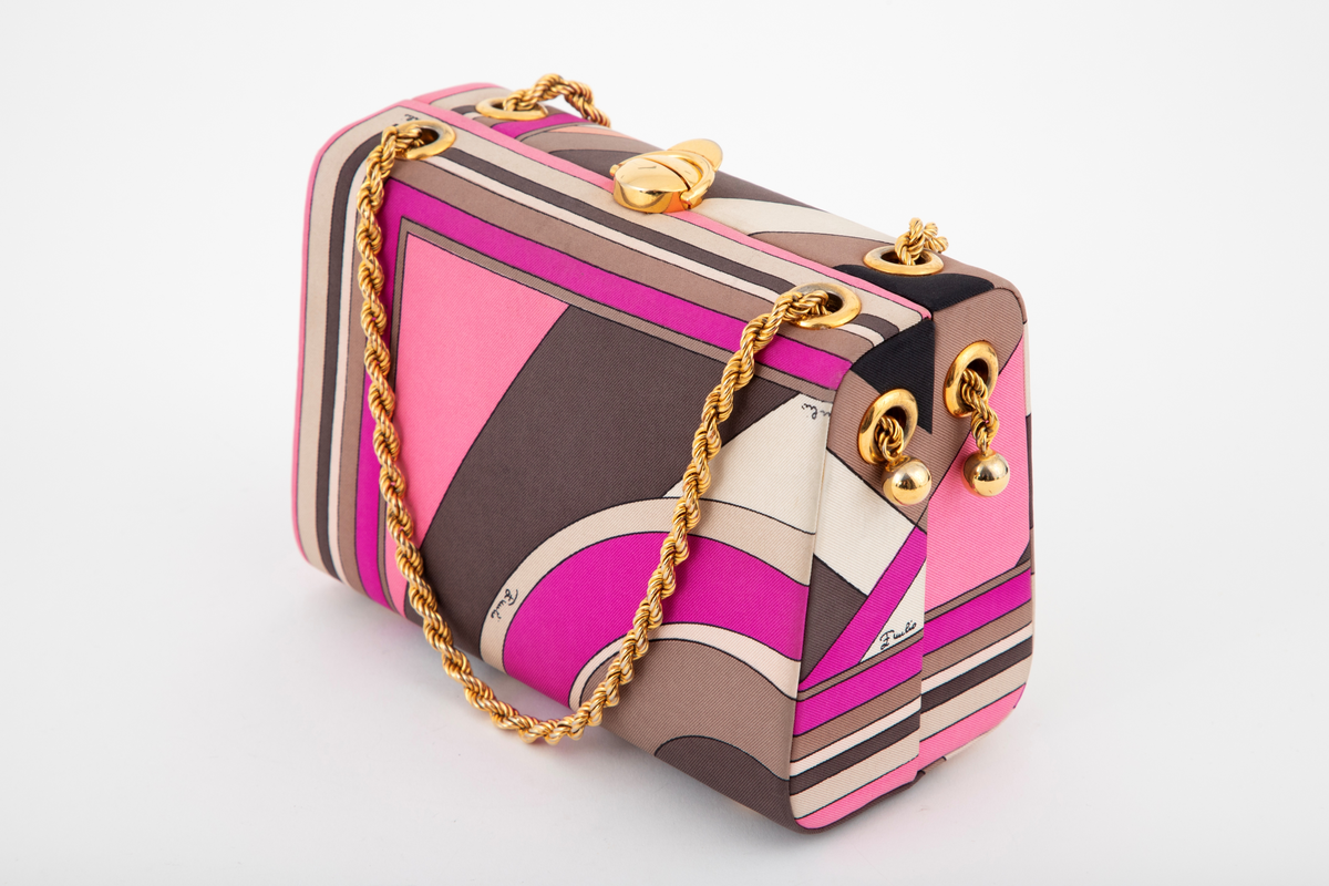 Liten boksveske med ett stort innerrom og et mindre innerrom på bakveggen. Vesken er hard, og må ha en innvendig kjerne av et hardt materiale. Utvendig er den trukket med et silketrykk i sterke rosa nyanser, nonfigurativt, men likevel noe geometrisk av type. Mønstertypen er karakteristisk for designeren Emilio Pucci på sluttet av 1960-tallet og på 1970-tallet. Innvendig kledd med hvitt kidskinn, og stor, båtformet åpning på innerlommen. Innerrommet er fôret med bronsefarget skinn. Knepplås på midten av oversiden og 2 bærelenker, tredd gjennom kraftige maljer på toppen og øverst på kortsidene. De tvunne "gullenkene" holdes på plass av små kuler i endene. På undersiden, 4 små halvkuleformede ben av samme metall.