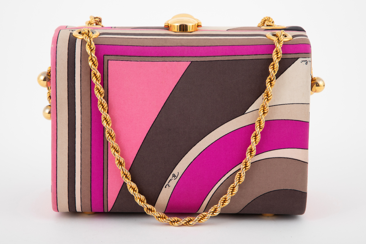 Liten boksveske med ett stort innerrom og et mindre innerrom på bakveggen. Vesken er hard, og må ha en innvendig kjerne av et hardt materiale. Utvendig er den trukket med et silketrykk i sterke rosa nyanser, nonfigurativt, men likevel noe geometrisk av type. Mønstertypen er karakteristisk for designeren Emilio Pucci på sluttet av 1960-tallet og på 1970-tallet. Innvendig kledd med hvitt kidskinn, og stor, båtformet åpning på innerlommen. Innerrommet er fôret med bronsefarget skinn. Knepplås på midten av oversiden og 2 bærelenker, tredd gjennom kraftige maljer på toppen og øverst på kortsidene. De tvunne "gullenkene" holdes på plass av små kuler i endene. På undersiden, 4 små halvkuleformede ben av samme metall.