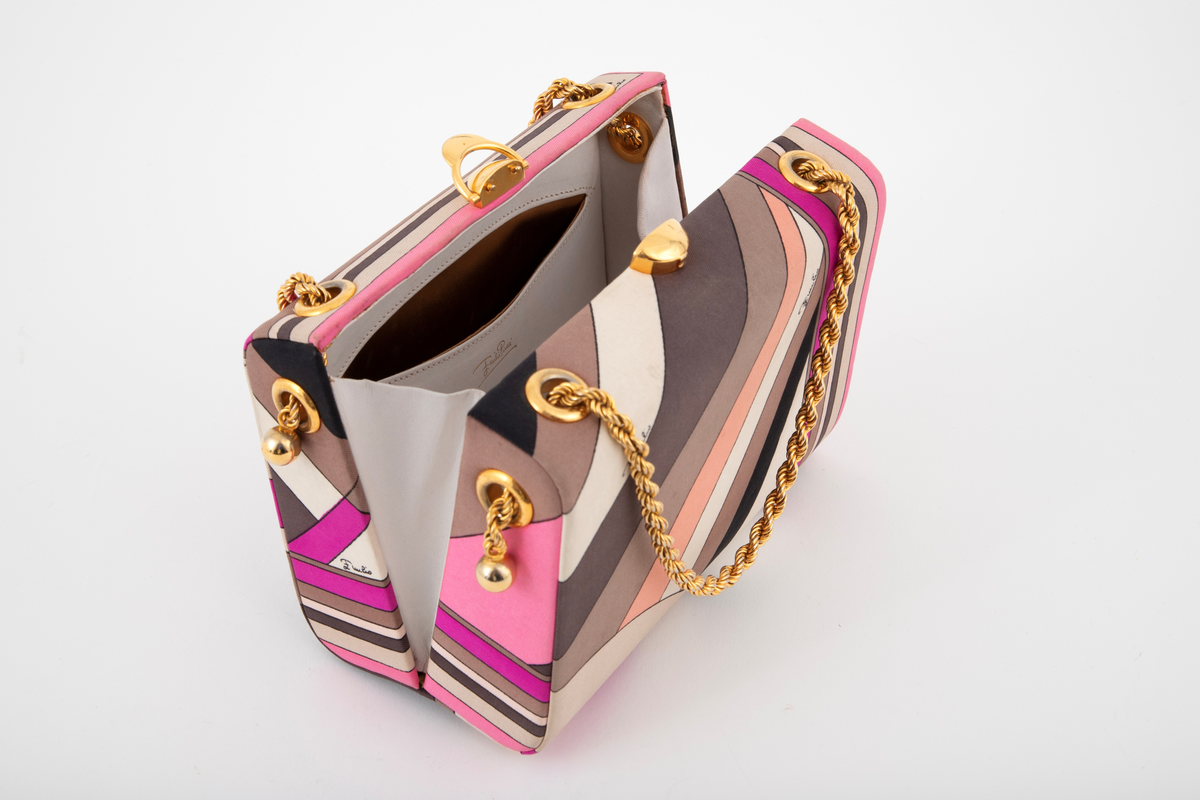 Liten boksveske med ett stort innerrom og et mindre innerrom på bakveggen. Vesken er hard, og må ha en innvendig kjerne av et hardt materiale. Utvendig er den trukket med et silketrykk i sterke rosa nyanser, nonfigurativt, men likevel noe geometrisk av type. Mønstertypen er karakteristisk for designeren Emilio Pucci på sluttet av 1960-tallet og på 1970-tallet. Innvendig kledd med hvitt kidskinn, og stor, båtformet åpning på innerlommen. Innerrommet er fôret med bronsefarget skinn. Knepplås på midten av oversiden og 2 bærelenker, tredd gjennom kraftige maljer på toppen og øverst på kortsidene. De tvunne "gullenkene" holdes på plass av små kuler i endene. På undersiden, 4 små halvkuleformede ben av samme metall.