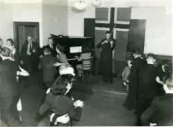 Dans i klasserommet ved Maskinistskulen i London under andre værdskrigen. Ein mann spelar fiolin, og ved pianoet sit ei kvinne og spelar. Det ligg to offisershattar på pianoet. Det norske flagget er hengt opp på veggen bak musikarane.