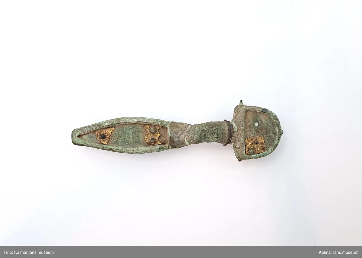 KLM 32783. Fibula. Gjuten av brons med pressbleck av guld samt delar som är försilvrade. Halvrunt överstycke med rester av pressbleck i guld fastsatt med två nitar. Runt kanten på överstycket finns spår av försilvring. Bågen har varit täckt med bleck, sannolikt av silver av vilket återstår endast en liten rest vid överstycket. Foten har haft pressbleck fastsatt med tre nitar och längs kanten finns spår av försilvring. Kort spiral och nål.