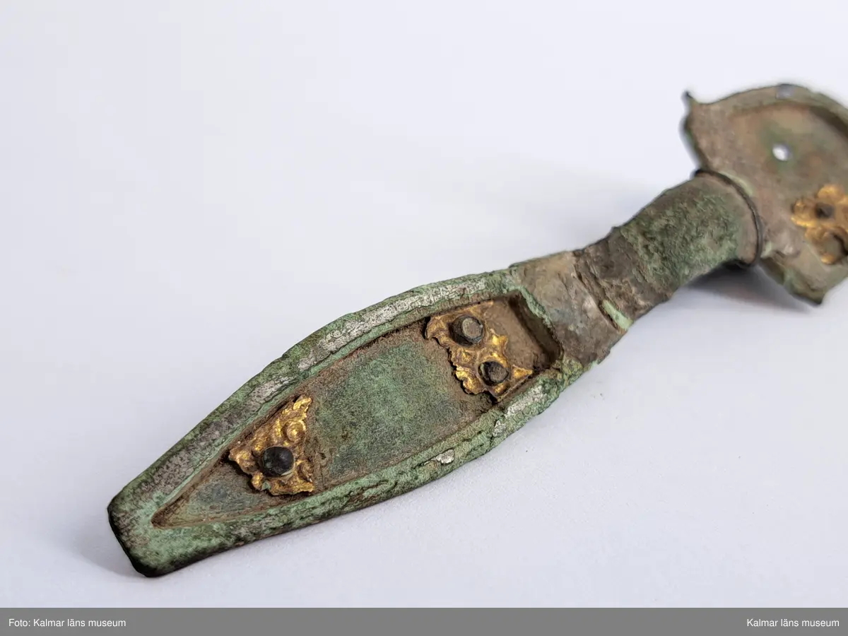 KLM 32783. Fibula. Gjuten av brons med pressbleck av guld samt delar som är försilvrade. Halvrunt överstycke med rester av pressbleck i guld fastsatt med två nitar. Runt kanten på överstycket finns spår av försilvring. Bågen har varit täckt med bleck, sannolikt av silver av vilket återstår endast en liten rest vid överstycket. Foten har haft pressbleck fastsatt med tre nitar och längs kanten finns spår av försilvring. Kort spiral och nål.