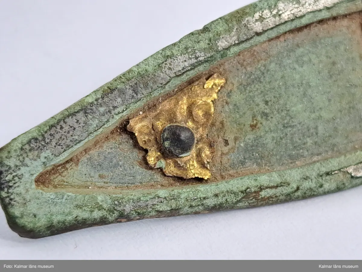 KLM 32783. Fibula. Gjuten av brons med pressbleck av guld samt delar som är försilvrade. Halvrunt överstycke med rester av pressbleck i guld fastsatt med två nitar. Runt kanten på överstycket finns spår av försilvring. Bågen har varit täckt med bleck, sannolikt av silver av vilket återstår endast en liten rest vid överstycket. Foten har haft pressbleck fastsatt med tre nitar och längs kanten finns spår av försilvring. Kort spiral och nål.