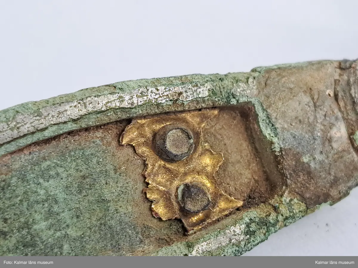 KLM 32783. Fibula. Gjuten av brons med pressbleck av guld samt delar som är försilvrade. Halvrunt överstycke med rester av pressbleck i guld fastsatt med två nitar. Runt kanten på överstycket finns spår av försilvring. Bågen har varit täckt med bleck, sannolikt av silver av vilket återstår endast en liten rest vid överstycket. Foten har haft pressbleck fastsatt med tre nitar och längs kanten finns spår av försilvring. Kort spiral och nål.