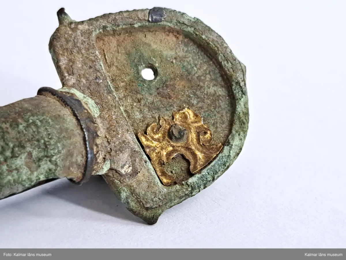 KLM 32783. Fibula. Gjuten av brons med pressbleck av guld samt delar som är försilvrade. Halvrunt överstycke med rester av pressbleck i guld fastsatt med två nitar. Runt kanten på överstycket finns spår av försilvring. Bågen har varit täckt med bleck, sannolikt av silver av vilket återstår endast en liten rest vid överstycket. Foten har haft pressbleck fastsatt med tre nitar och längs kanten finns spår av försilvring. Kort spiral och nål.