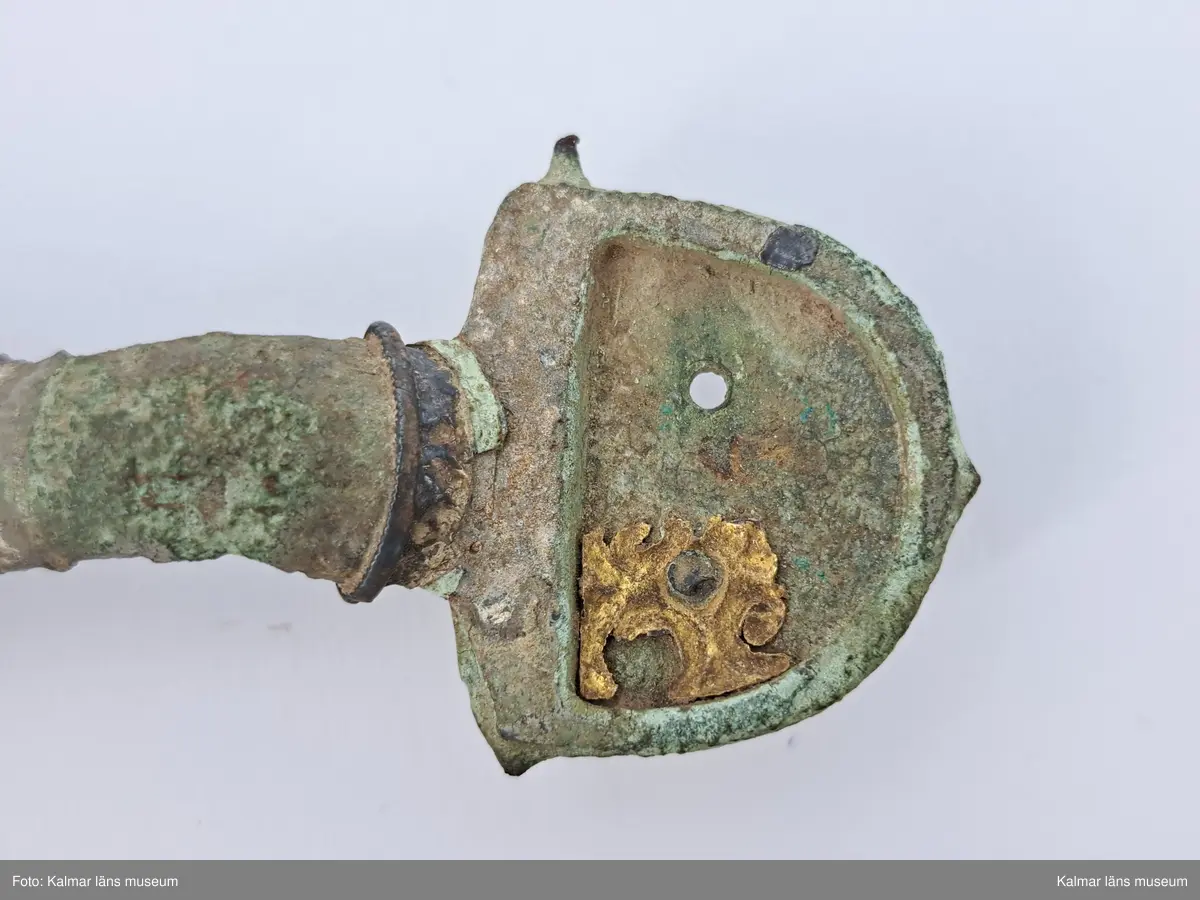 KLM 32783. Fibula. Gjuten av brons med pressbleck av guld samt delar som är försilvrade. Halvrunt överstycke med rester av pressbleck i guld fastsatt med två nitar. Runt kanten på överstycket finns spår av försilvring. Bågen har varit täckt med bleck, sannolikt av silver av vilket återstår endast en liten rest vid överstycket. Foten har haft pressbleck fastsatt med tre nitar och längs kanten finns spår av försilvring. Kort spiral och nål.