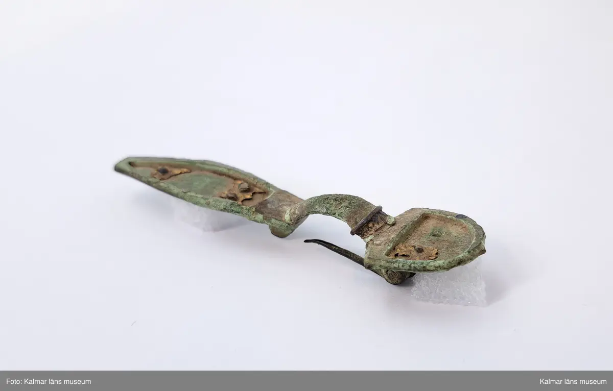 KLM 32783. Fibula. Gjuten av brons med pressbleck av guld samt delar som är försilvrade. Halvrunt överstycke med rester av pressbleck i guld fastsatt med två nitar. Runt kanten på överstycket finns spår av försilvring. Bågen har varit täckt med bleck, sannolikt av silver av vilket återstår endast en liten rest vid överstycket. Foten har haft pressbleck fastsatt med tre nitar och längs kanten finns spår av försilvring. Kort spiral och nål.