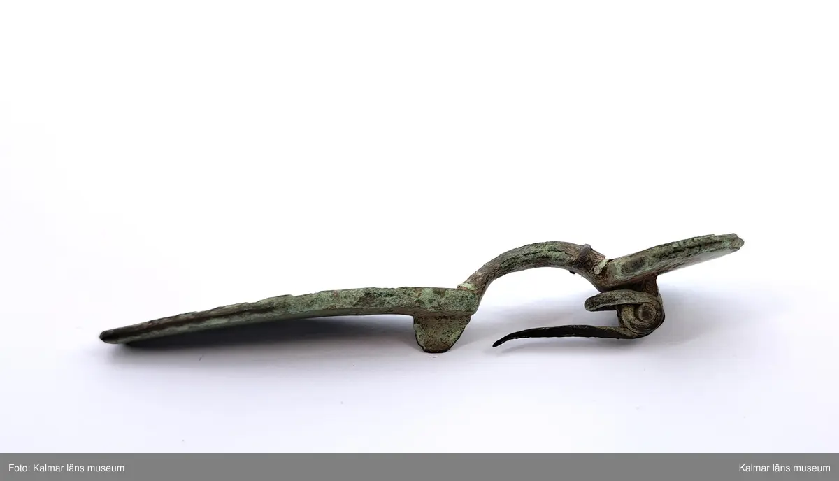 KLM 32783. Fibula. Gjuten av brons med pressbleck av guld samt delar som är försilvrade. Halvrunt överstycke med rester av pressbleck i guld fastsatt med två nitar. Runt kanten på överstycket finns spår av försilvring. Bågen har varit täckt med bleck, sannolikt av silver av vilket återstår endast en liten rest vid överstycket. Foten har haft pressbleck fastsatt med tre nitar och längs kanten finns spår av försilvring. Kort spiral och nål.