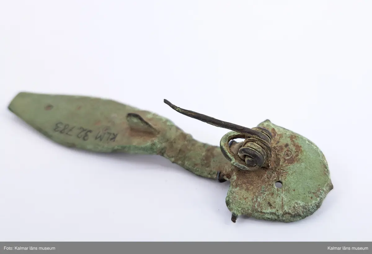 KLM 32783. Fibula. Gjuten av brons med pressbleck av guld samt delar som är försilvrade. Halvrunt överstycke med rester av pressbleck i guld fastsatt med två nitar. Runt kanten på överstycket finns spår av försilvring. Bågen har varit täckt med bleck, sannolikt av silver av vilket återstår endast en liten rest vid överstycket. Foten har haft pressbleck fastsatt med tre nitar och längs kanten finns spår av försilvring. Kort spiral och nål.