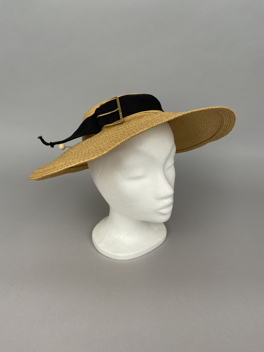 Hatt av tunn flätad ofärgad strå- eller remsa av annan växtfiber. Låg smal kulle omsvept av svart hattband i sidenrips. Bandet går ej runt hela kullen och har ett spänne av gulmetall. Modell som ska sitta ovanpå huvudet fastsatt med en nål. Brättet är vidare fram än bak.