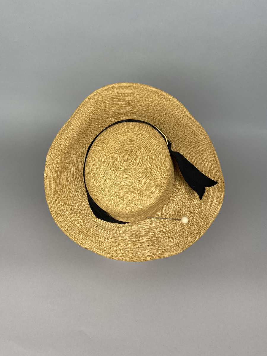 Hatt av tunn flätad ofärgad strå- eller remsa av annan växtfiber. Låg smal kulle omsvept av svart hattband i sidenrips. Bandet går ej runt hela kullen och har ett spänne av gulmetall. Modell som ska sitta ovanpå huvudet fastsatt med en nål. Brättet är vidare fram än bak.