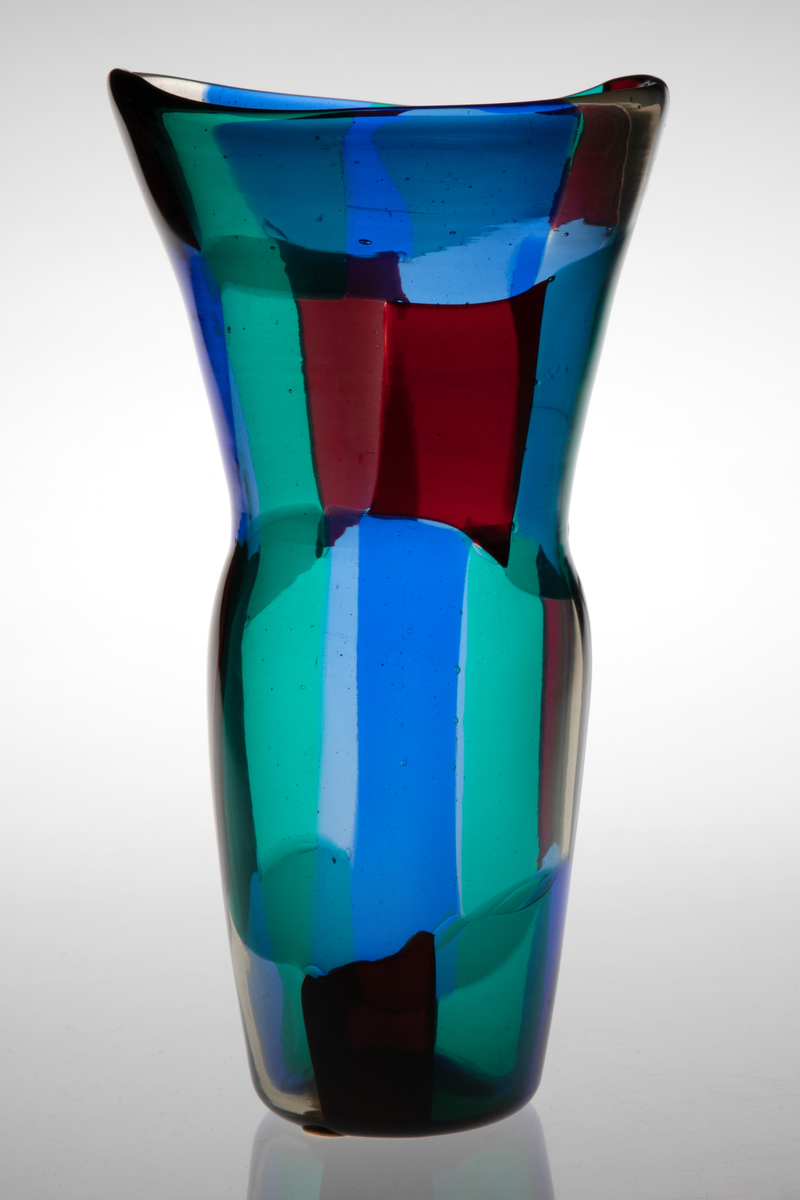 Vase i gjennomskinnelig glass. Korpus er dekorert med rektangulære felter i grønt, rødt, blått og klart glass. Ujevn ovalformet munning og lang konisk hals.