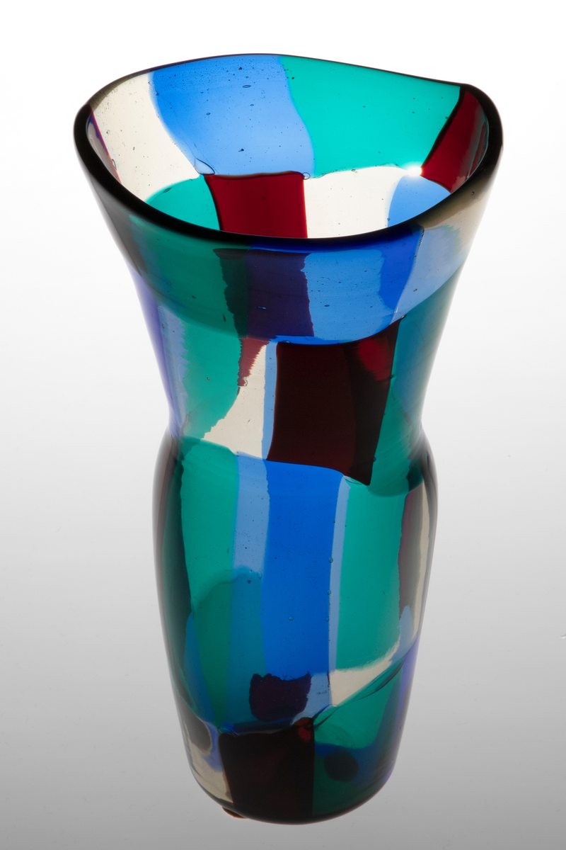 Vase i gjennomskinnelig glass. Korpus er dekorert med rektangulære felter i grønt, rødt, blått og klart glass. Ujevn ovalformet munning og lang konisk hals.