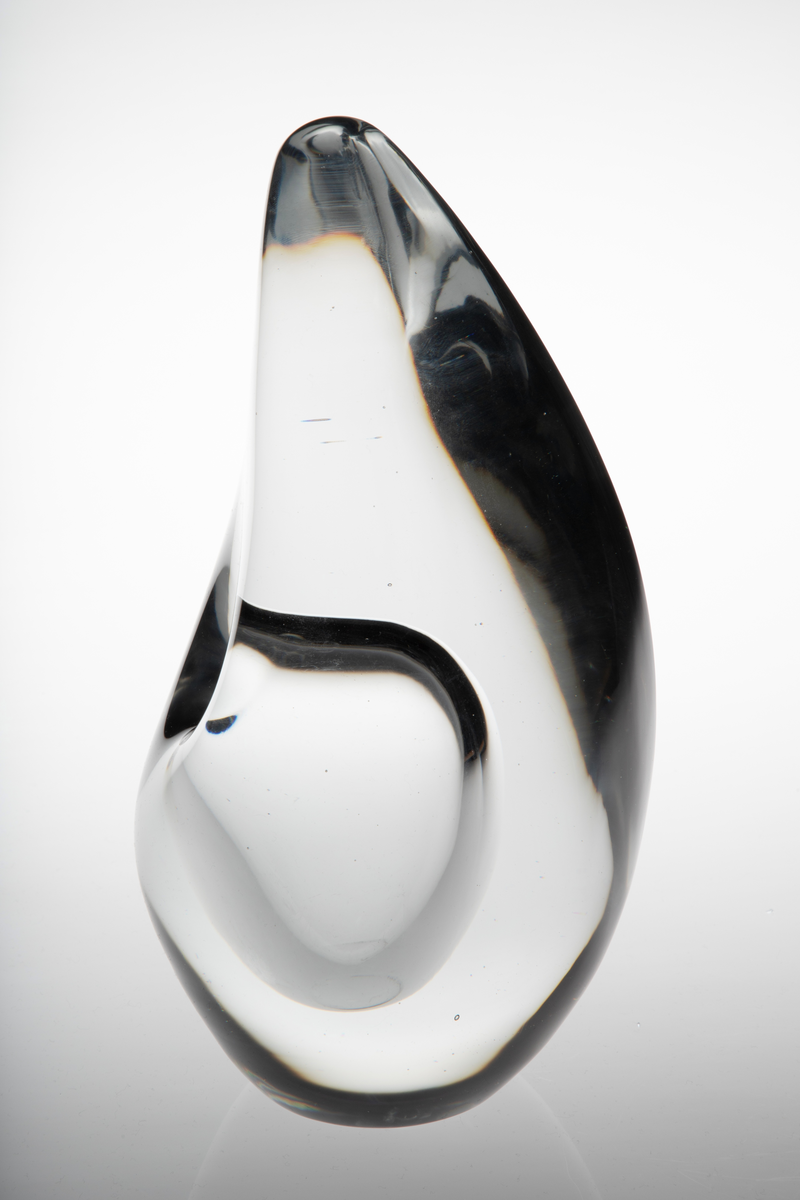 Dråpeformet skulpturell vase med abstrakt utforming i massivt klart glass. Et ellipseformet hulerom i nedre del med liten sirkulær åpning. Bunnen er planslipt.