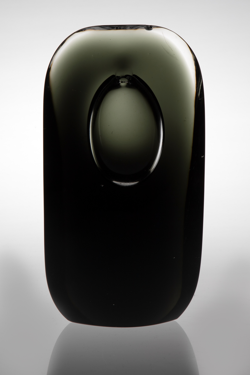 Skulpturell vase i klart og farget glass, hvor sistnevnte er opakt med et mørkt grå-grønnlig skjær. Rektangulær hovedform med mykt avrundede hjørner samt svakt konveks for- og bakside. På øvre del en ellipseformet luftboble i klart glass med en liten åpning. Planslipt bunn.