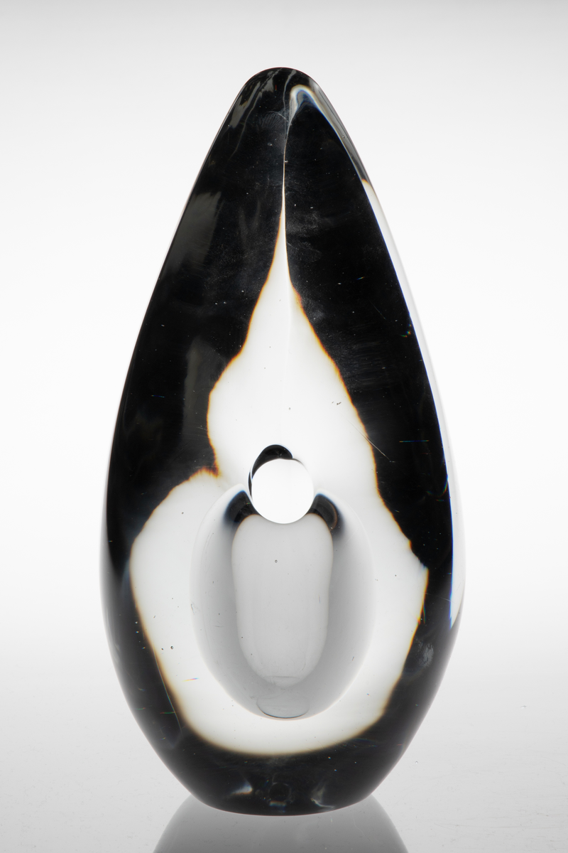 Dråpeformet skulpturell vase med abstrakt utforming i massivt klart glass. Et ellipseformet hulerom i nedre del med liten sirkulær åpning. Bunnen er planslipt.