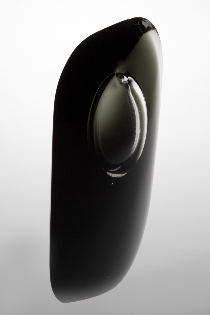 Skulpturell vase i klart og farget glass, hvor sistnevnte er opakt med et mørkt grå-grønnlig skjær. Rektangulær hovedform med mykt avrundede hjørner samt svakt konveks for- og bakside. På øvre del en ellipseformet luftboble i klart glass med en liten åpning. Planslipt bunn.