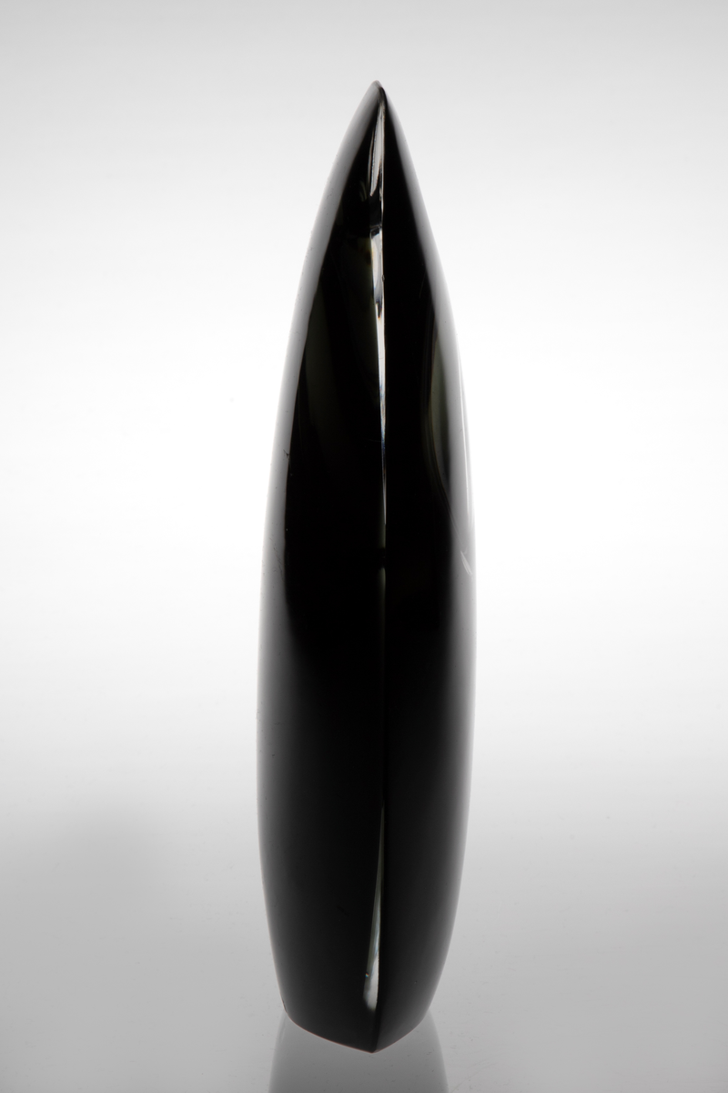 Skulpturell vase i klart og farget glass, hvor sistnevnte er opakt med et mørkt grå-grønnlig skjær. Rektangulær hovedform med mykt avrundede hjørner samt svakt konveks for- og bakside. På øvre del en ellipseformet luftboble i klart glass med en liten åpning. Planslipt bunn.