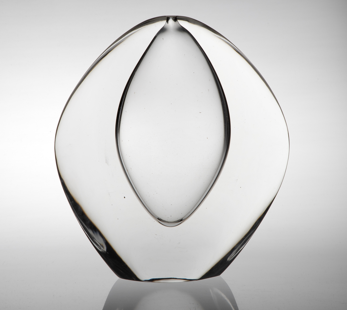 Vase av klart glass med skulpturell utforming. Flattrykket oval hovedform med konvekse sider. Ellipseformet hulerom i midten med en liten åpning på toppen.
